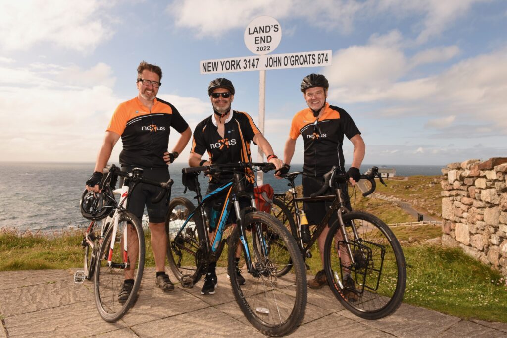 Three cyclists in black and orange Nexus jerseys stand with their bikes at the iconic Land’s End signpost, marking the finish of a long-distance ride
