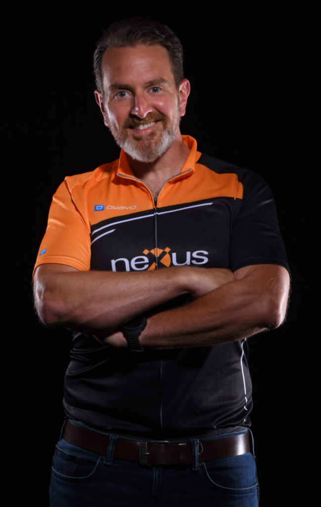 Portrait of a smiling man in a black and orange Nexus cycling jersey standing with arms crossed against a dark background
