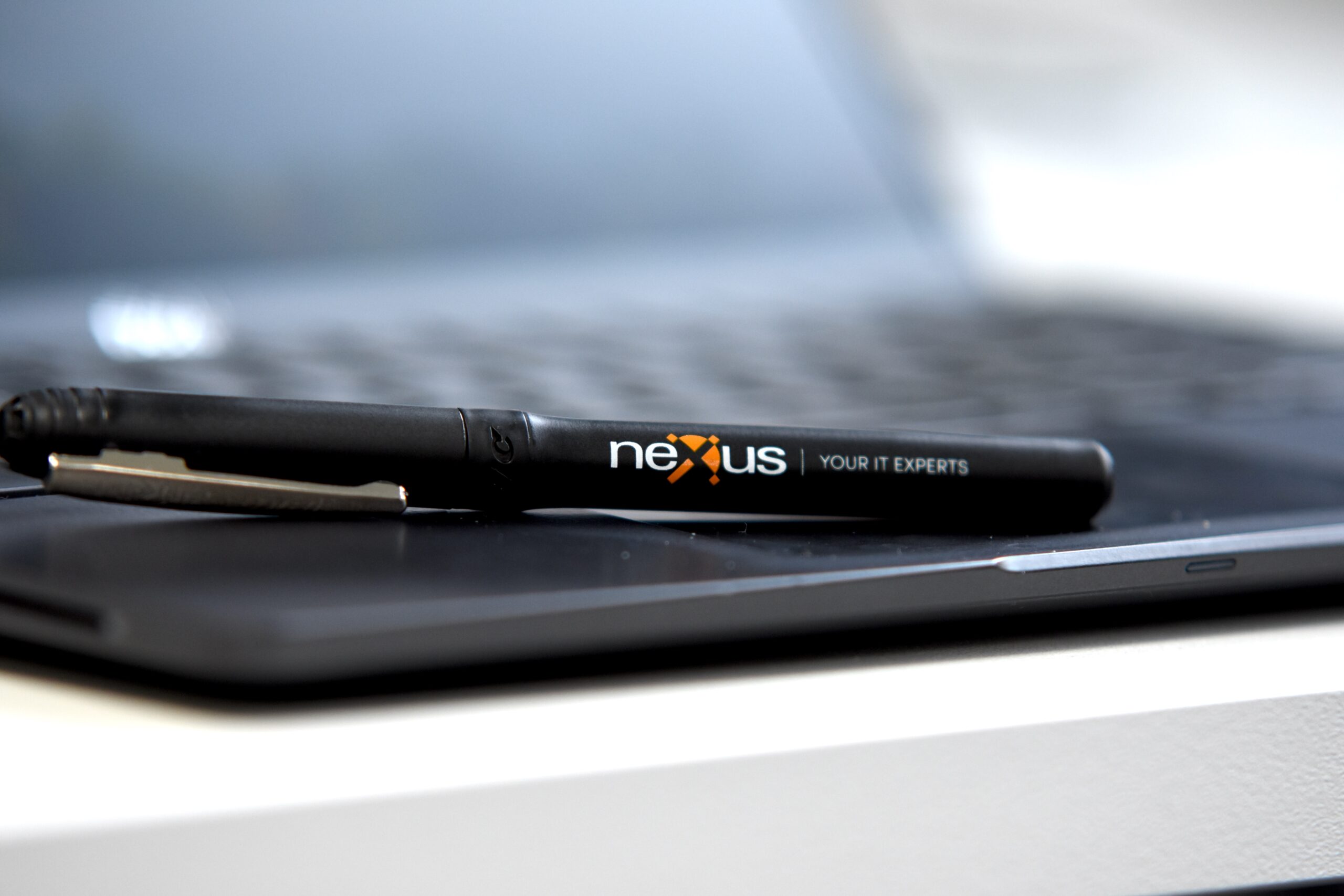 Close-up of a black pen branded with the Nexus logo and tagline “Your IT Experts,” resting on a laptop keyboard