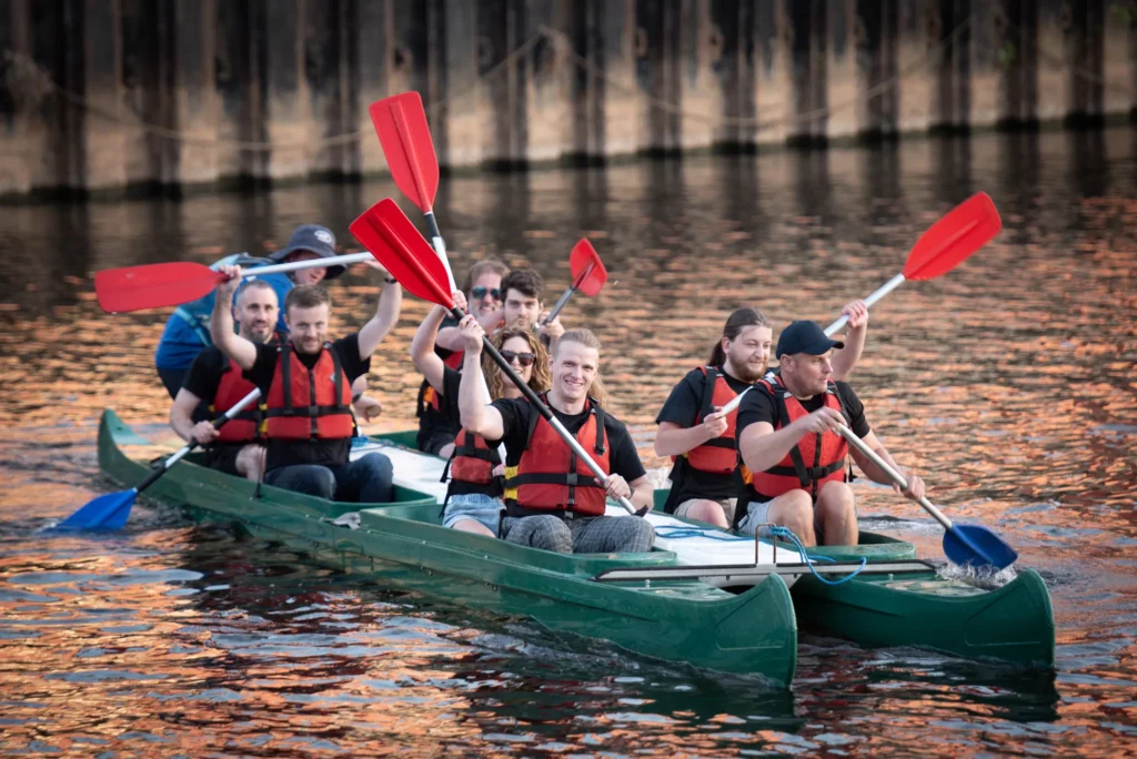 Nexus team wearing life jackets paddling and smiling in a green canoe on a calm body of water during a team-building activity.