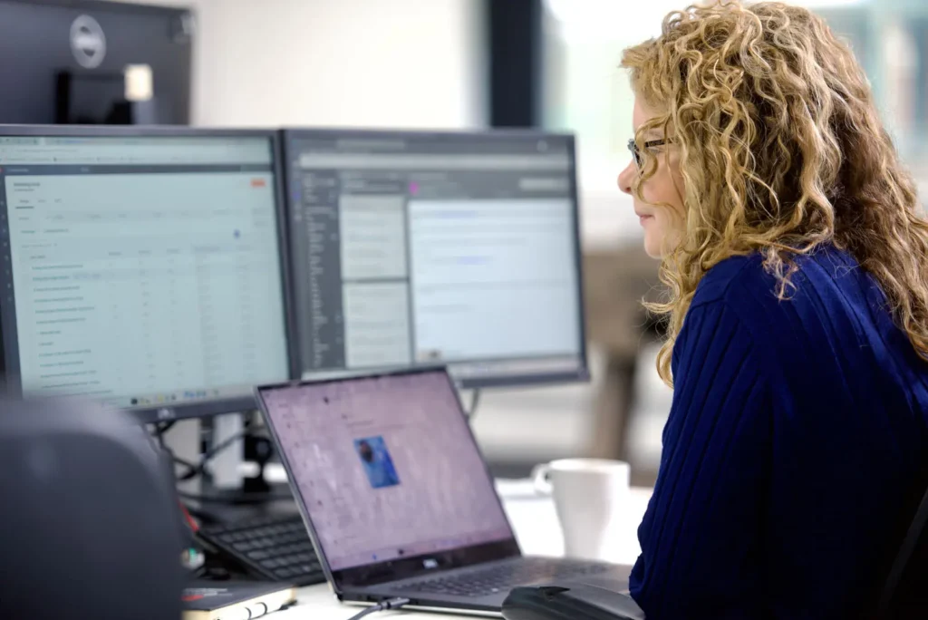 Woman in glasses working on detailed data across several monitors in an office environment