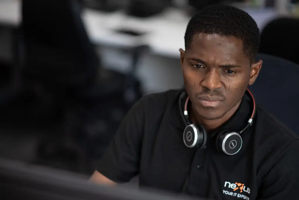 Man wearing a black Nexus IT Experts polo shirt and headphones around his neck, focused on a computer screen in an office
