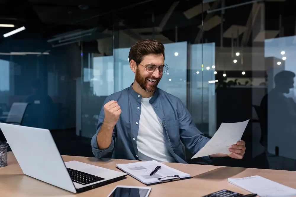 A happy young man, a businessman, a freelancer sits in the office, looking and holds a document.