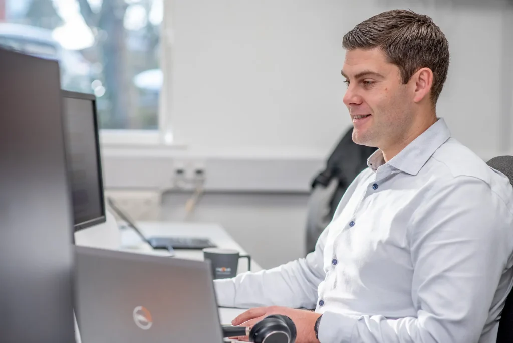 Nexus IT Experts team member in a white shirt smiling while working at a laptop and monitor in the office