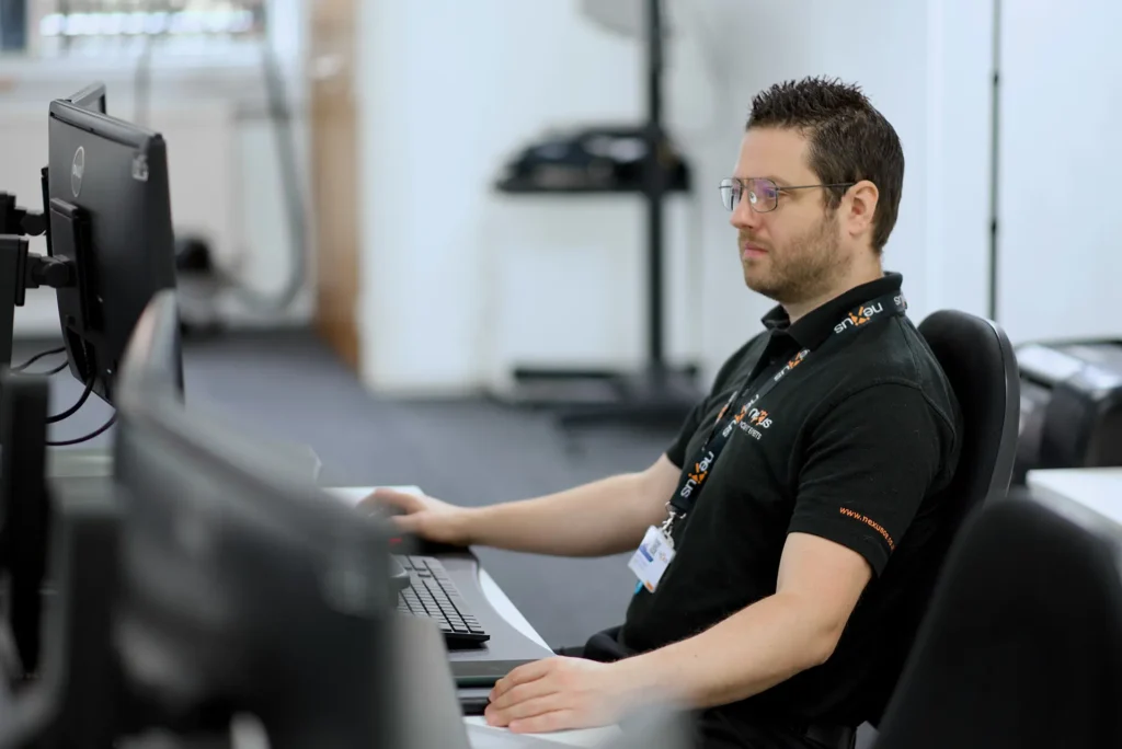 Nexus IT Experts team member in a black polo shirt and lanyard working at a computer workstation in the office