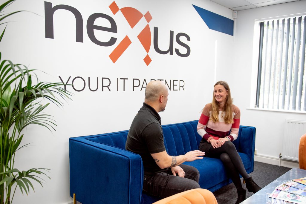 Two colleagues chatting on a blue velvet sofa in a bright office, beneath a large Nexus "Your IT Partner" logo on the wall